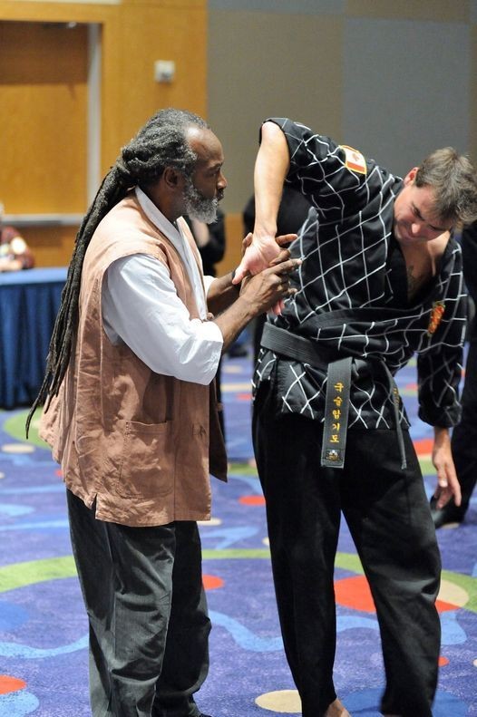 Two individuals practicing a martial arts technique in a room with colorful carpet and wooden walls.